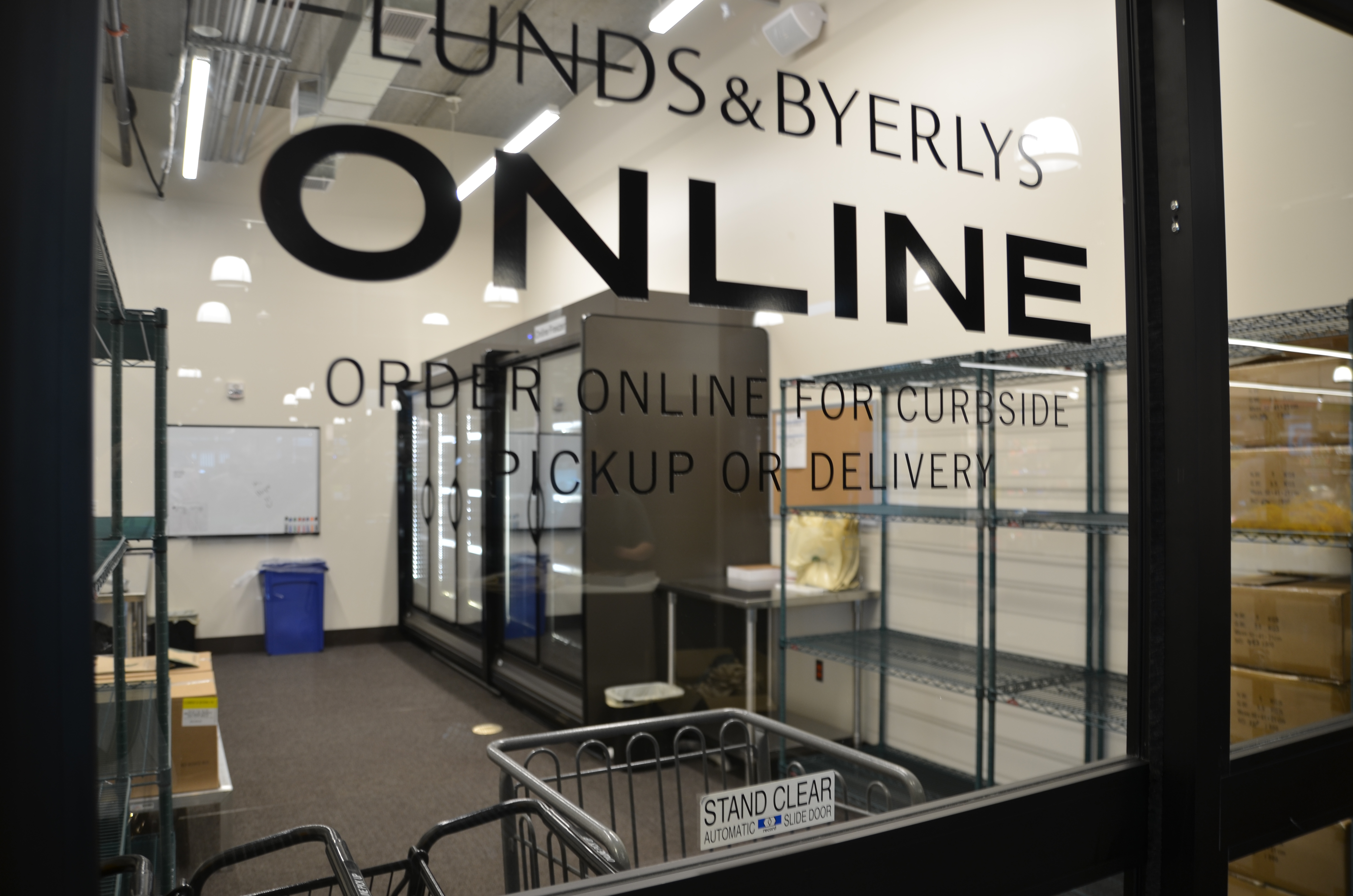 A room with shelves and refrigerators. Lunds and Byerlys online, order online for curbside pickup or delivery is written on the window.